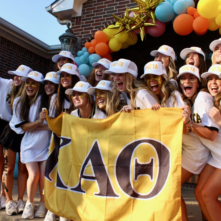 Group of CPC members smiling while wearing hats that say “Theta” and posing with a banner displaying their Greek letters for their chapter